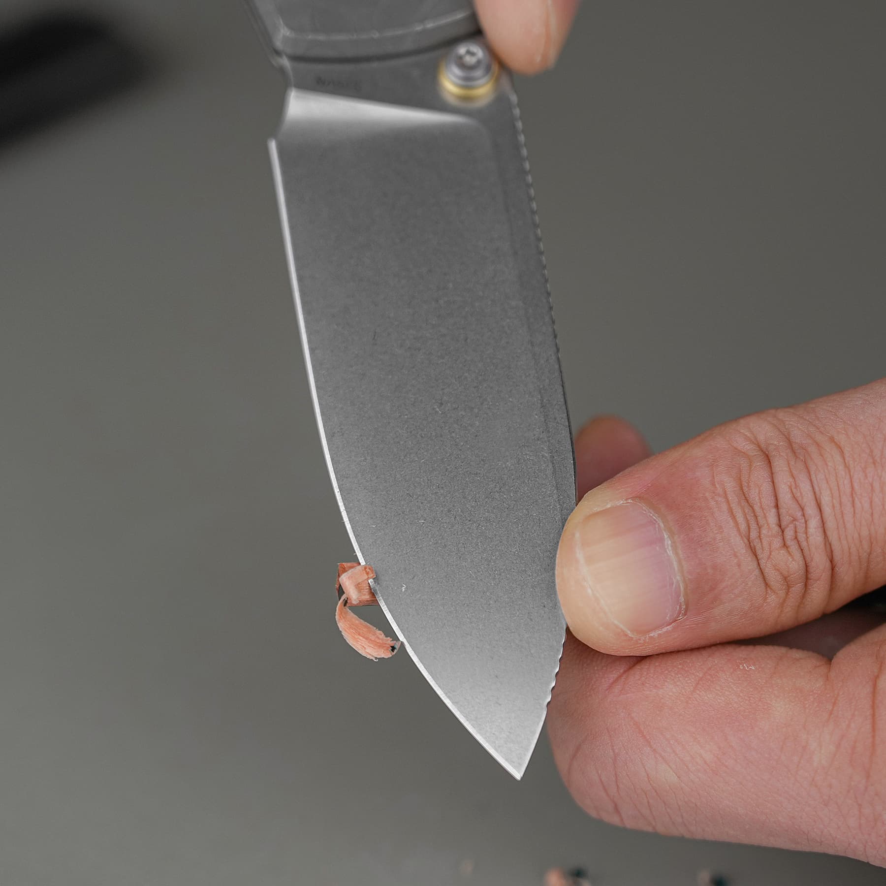 Close-up of a hand holding a curved metal blade, with a small copper strip curling off its edge during sharpening.