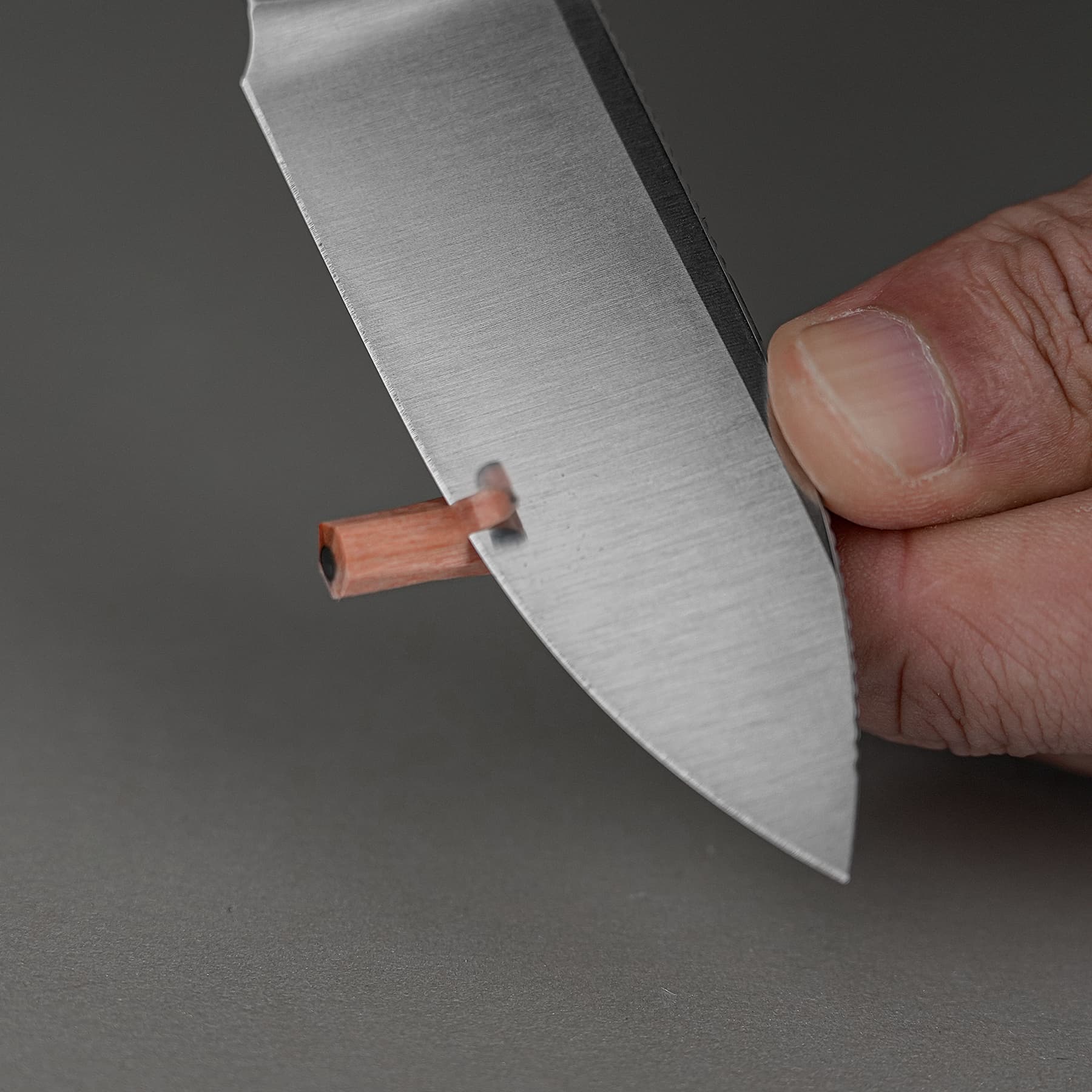 Close-up of a hand holding a straight blade with a small orange-copper rectangular piece at the tip against a gray background.