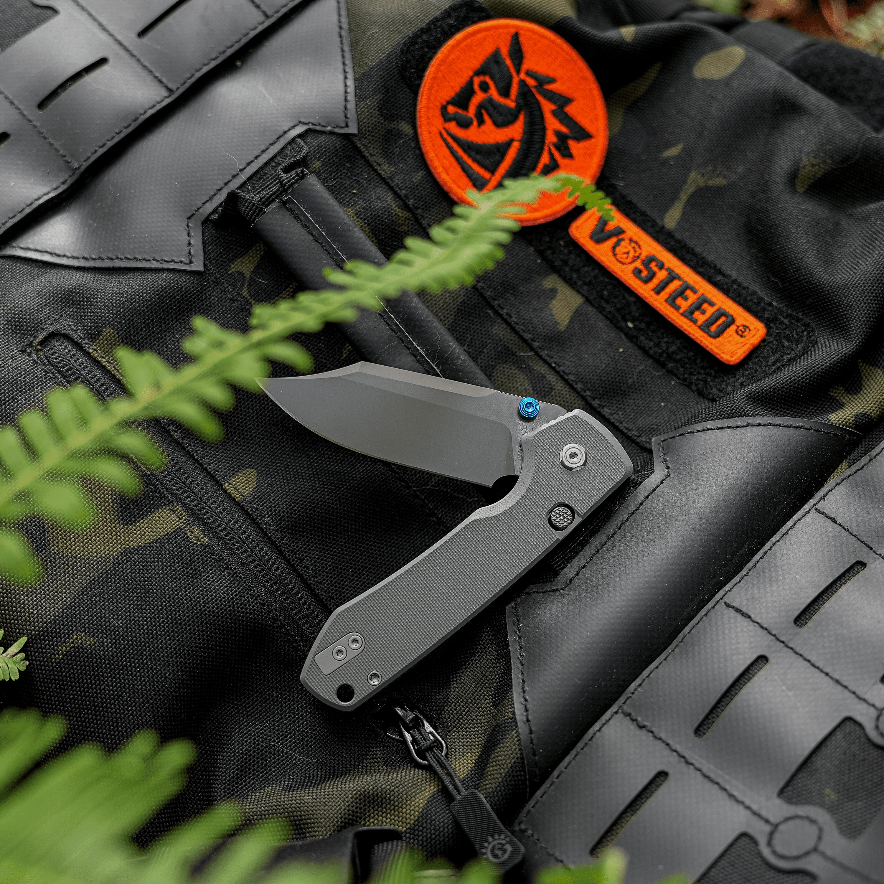 Close-up of camouflage tactical vest with an orange circular patch and a folding knife nearby on black gear fabric