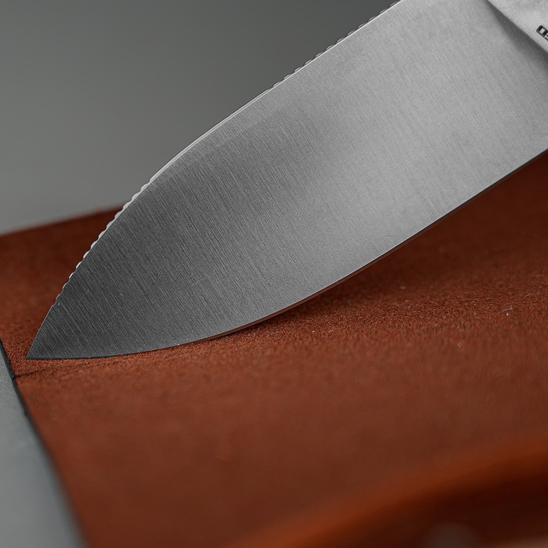 Close-up of a sharp metal knife blade with fine horizontal grain and a serrated edge resting on a reddish-brown surface