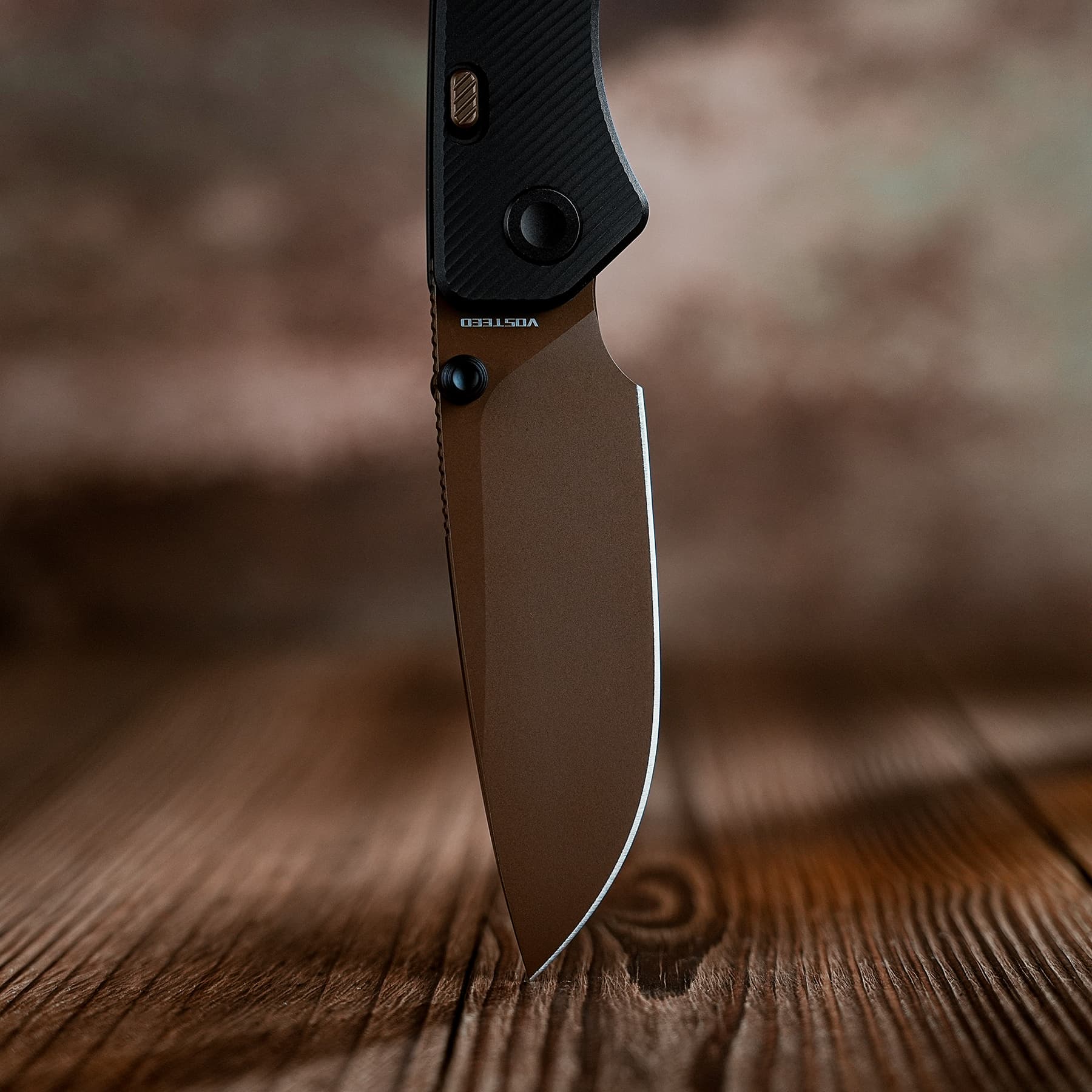 Close-up of a folding knife blade tip piercing a wooden tabletop, with a textured black handle above
