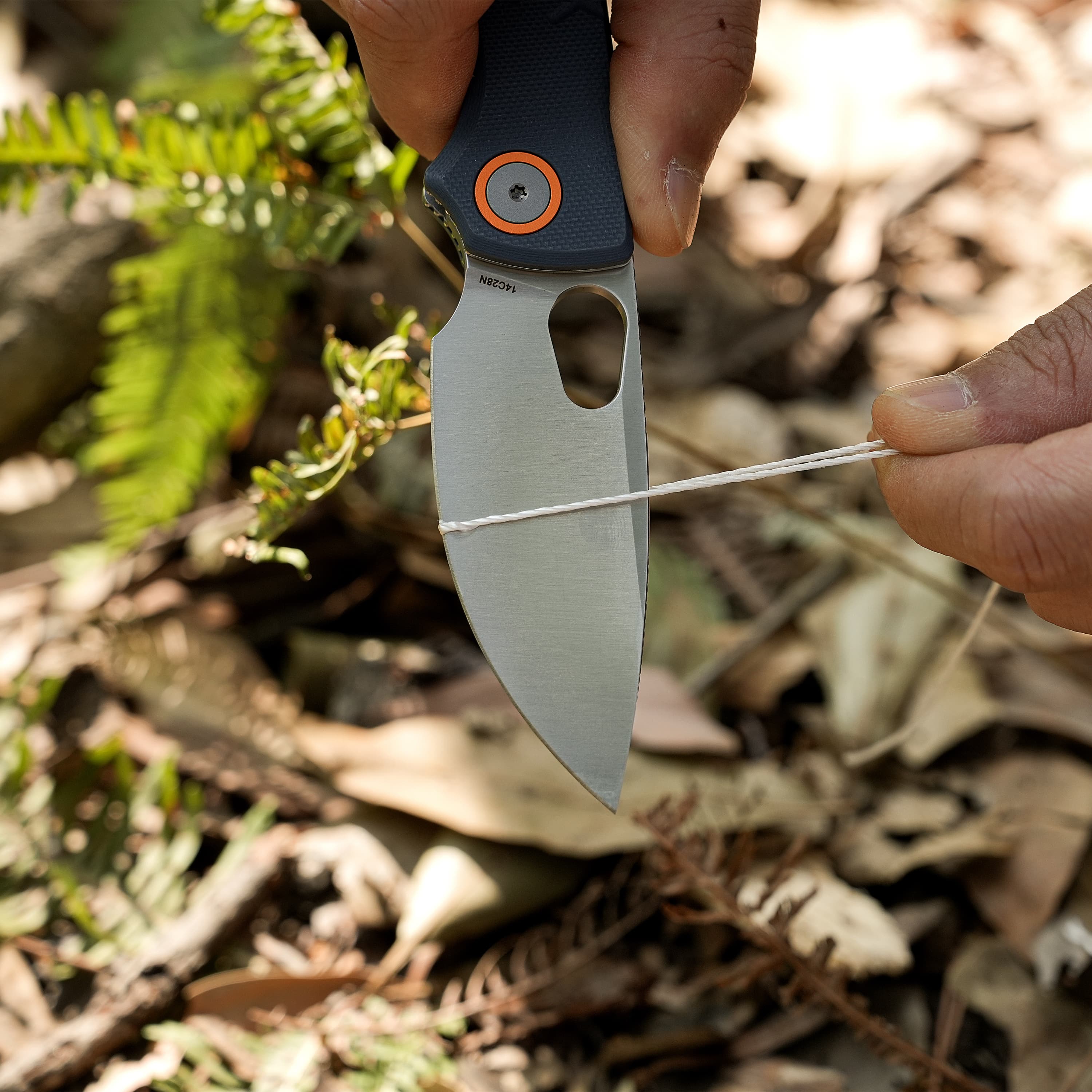 Close-up of a folding knife blade held by fingers, showing an orange-ringed pivot screw on a dark textured handle outdoors.