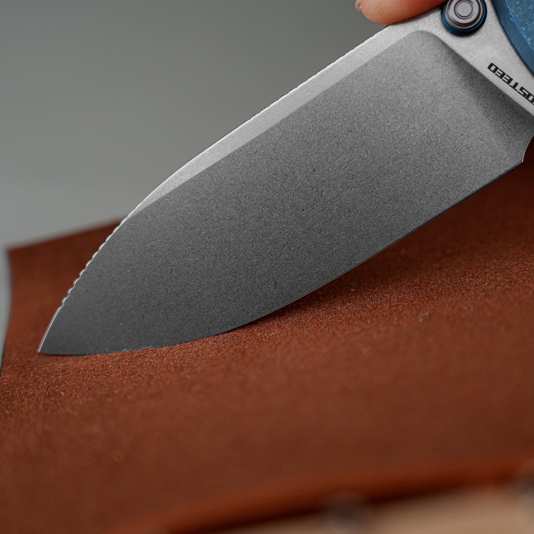 Close-up of a gray serrated knife blade resting on a reddish-brown surface, with part of the handle and a blue fastener visible.
