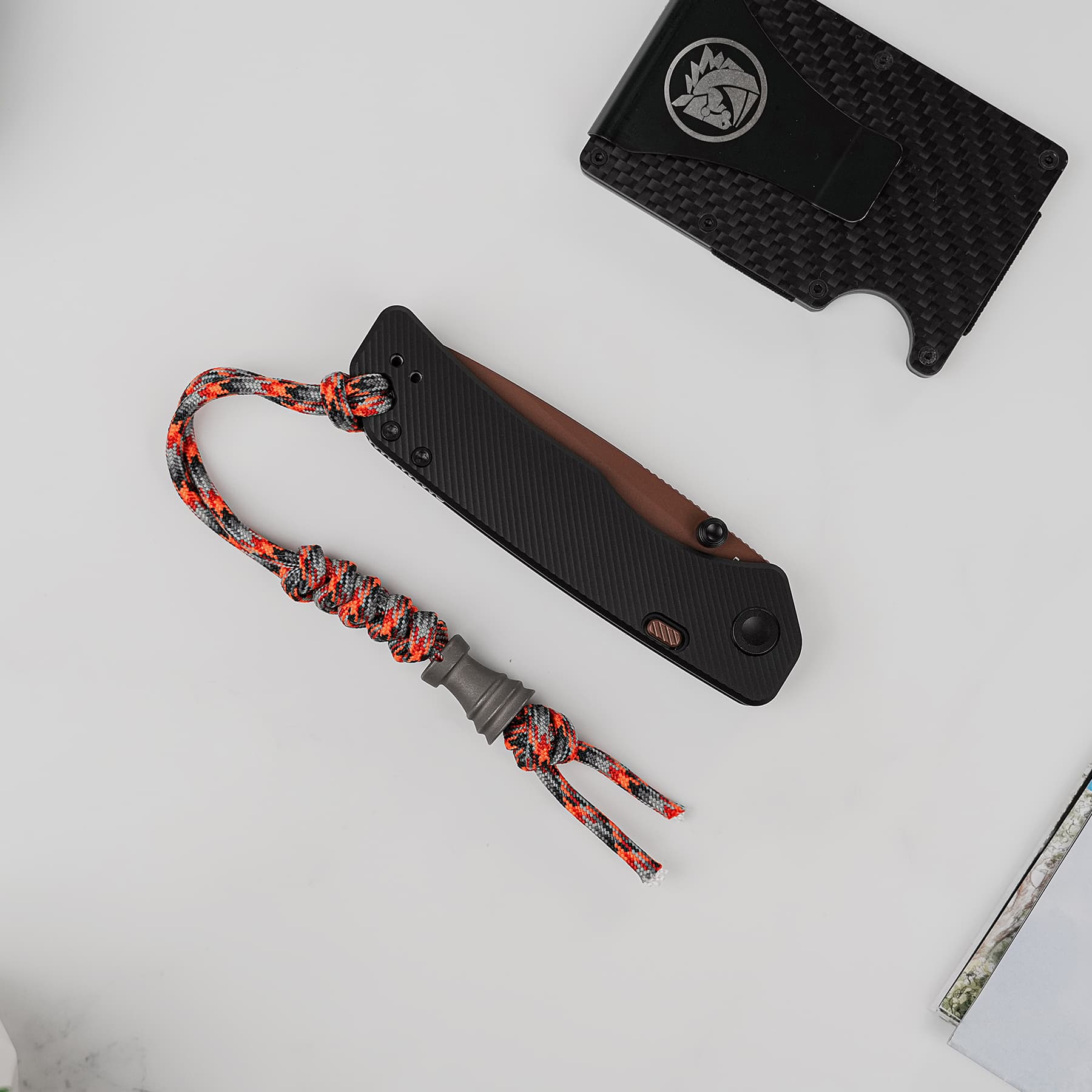 Close-up of a black folding knife with a textured handle and an orange-gray paracord lanyard on a light gray surface, with part of a black sheath nearby.
