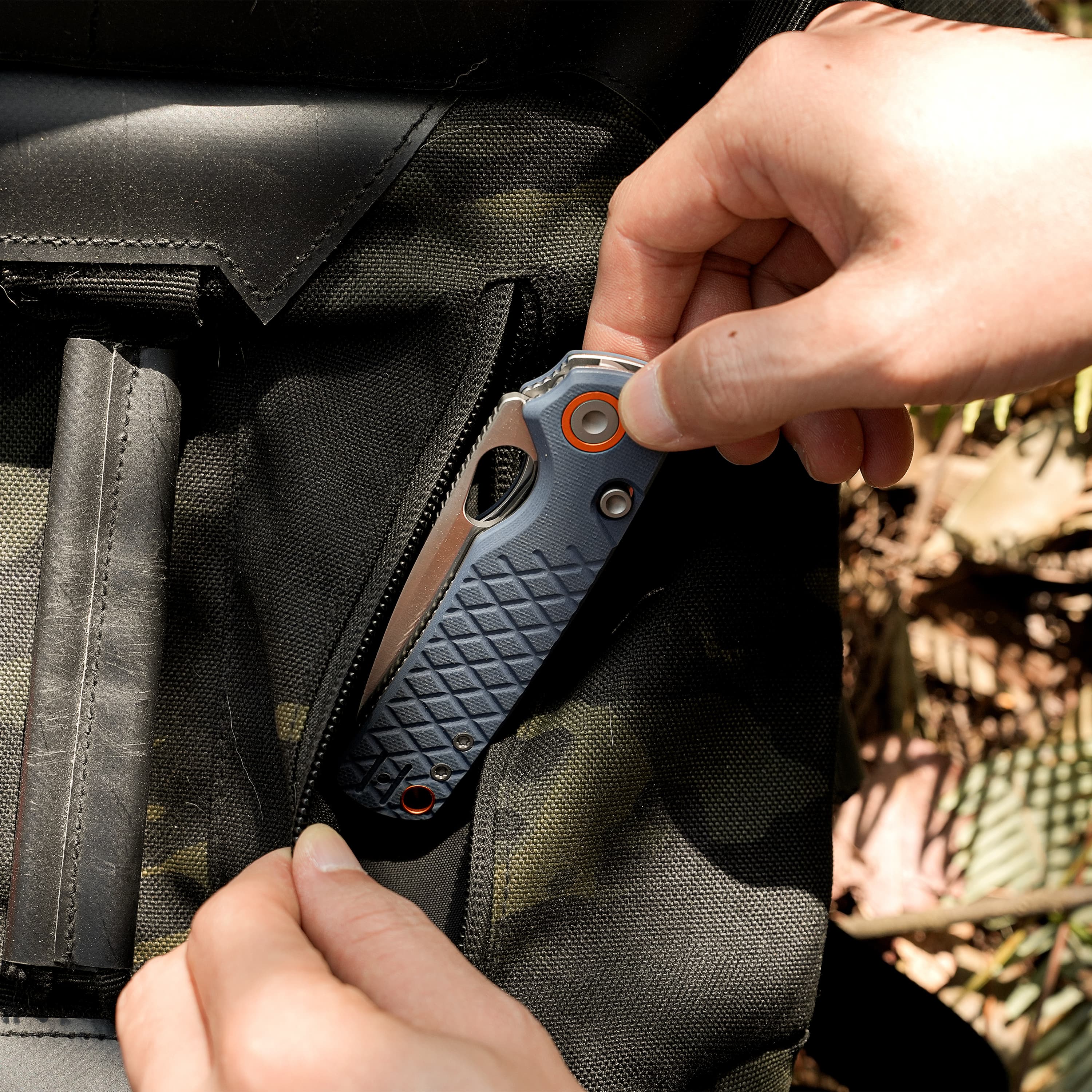 Close-up of a hand unzipping a dark fabric backpack, exposing a gray multitool with orange accents inside.