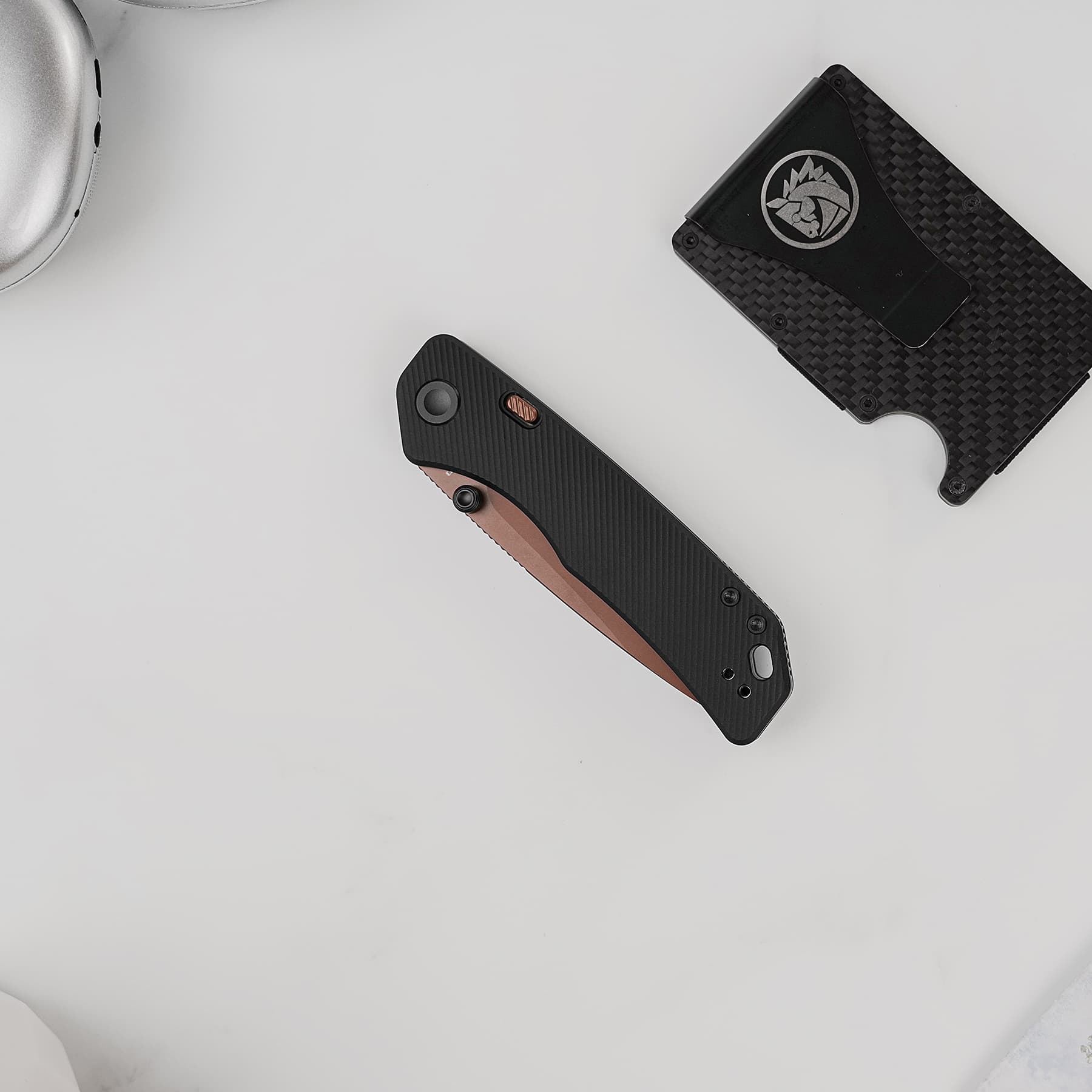 Two folding knives with textured handles placed on a pale gray surface; one has a reddish-brown blade edge, the other features a black carbon-fiber pattern and a circular logo.