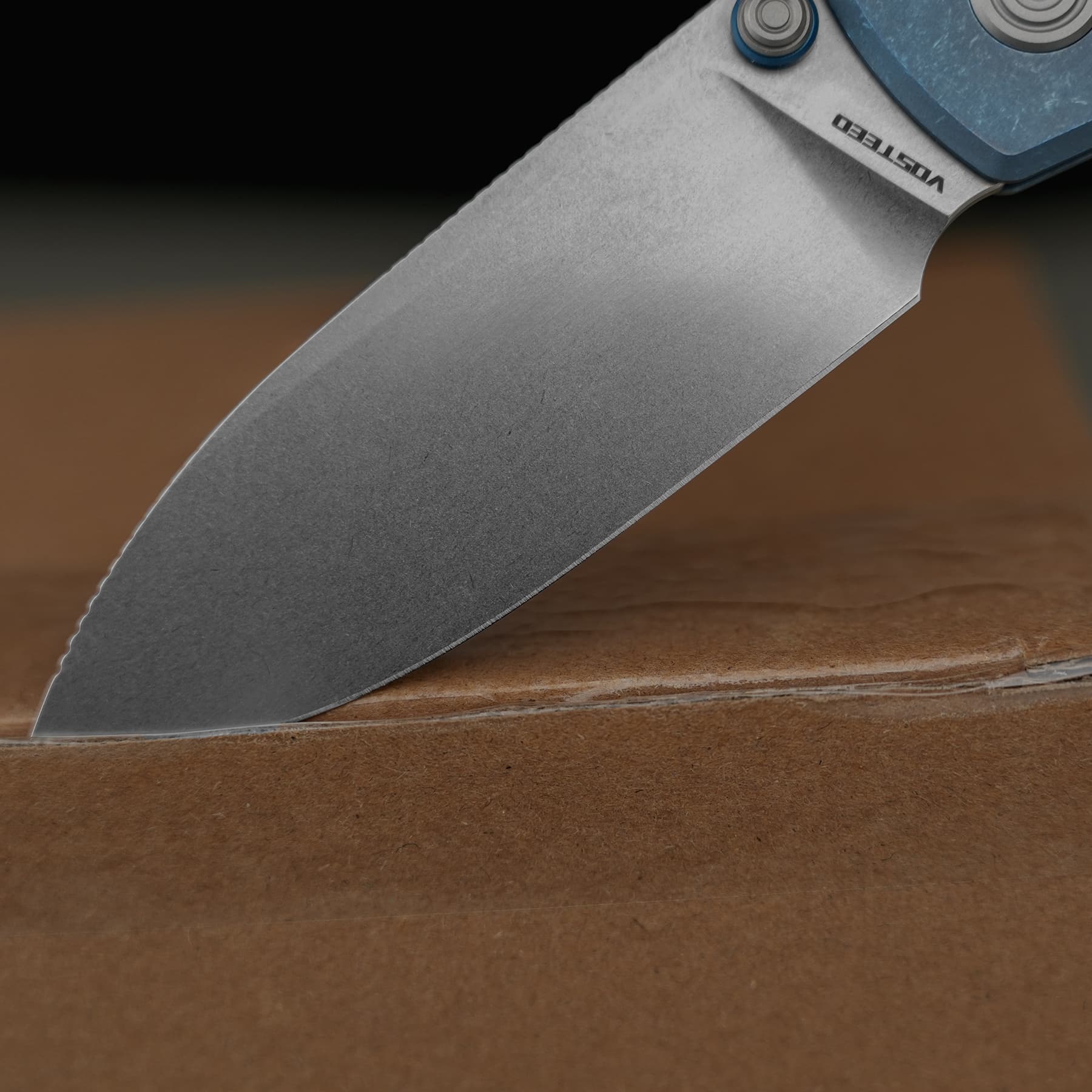 Close-up of a metal folding knife blade resting on a wooden surface, with a blue handle visible at the top right
